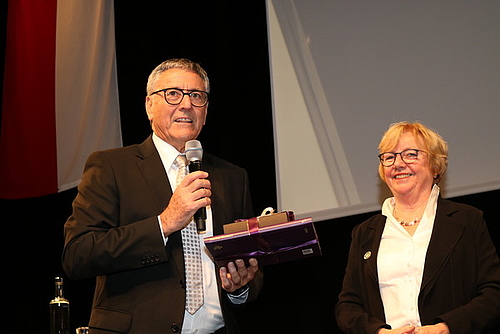 Bürgermeister Wolfgang Matt hält in einer Hand ein Mikrofon, in das er spricht. In der anderen Hand hält er ein Geschenk für die Rednerin Christel Bienstein, welche neben ihm steht.