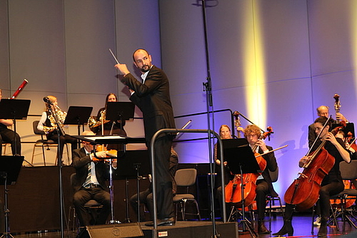 Ein Dirigent steht vor dem Orchester, er blickt über seine Schulter.