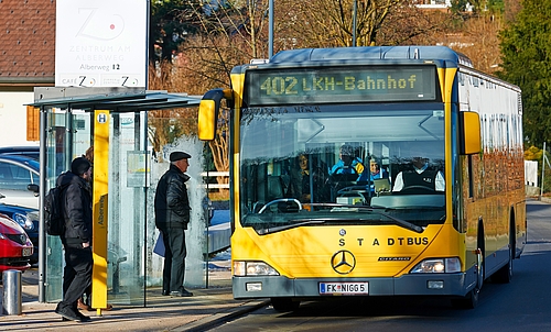 Ein Stadtbus hält an einer Haltestelle, an der drei Männer stehen. 