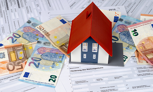 Mehrere Geldscheine und Dokumente liegen durcheinander, darauf steht ein Modell eines Hauses.