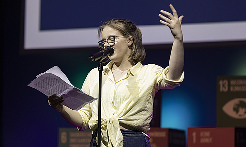 Ein junges Mädchen steht auf der Bühne und spricht in ein Mikrofon, das vor ihr steht. In einer Hand hält sie Zettel, von denen sie abliest. Die andere Hand hält sie nach oben.