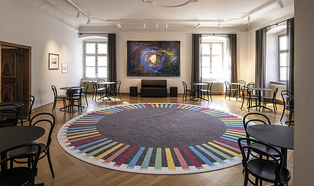 Raum im Palais Liechtenstein, ein großer runder Teppich mit bunten Streifen, um den Teppich herum stehen schwarze Tische und Stühle.