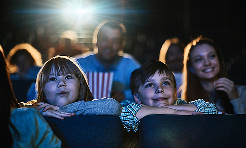 Mehrere Personen sitzen in einem Kinosaal und schauen einen Film. Zwei Kindder haben ihre Köpfe auf den Sitzen vor ihnen aufgelegt.