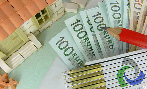 Ein Modell eines Hauses, ein Bleistift, ein Meterstab und Geldscheine liegen auf einem Tisch. 