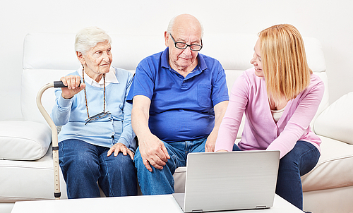 Ein älteres Paar sitzt auf einer Couch, neben ihnen sitzt eine jüngere Frau, die ihnen etwas auf dem Laptop zeigt.