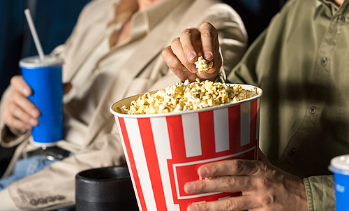 Ein älterer Mann und eine ältere Frau sitzen in einem Kinosaal. Der Mann hält einen Becher Popcorn in den Händen und die Frau einen Getränkebecher. 