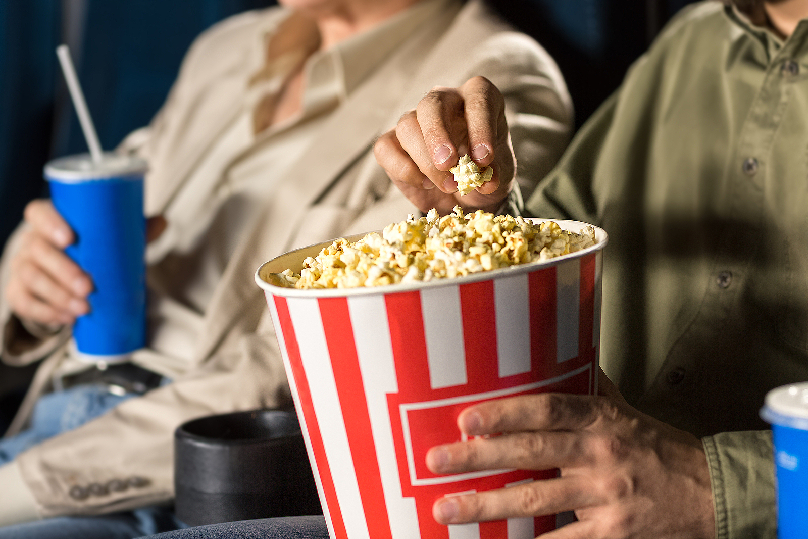 Ein älterer Mann und eine ältere Frau sitzen in einem Kinosaal. Der Mann hält einen Becher Popcorn in den Händen und die Frau einen Getränkebecher. 