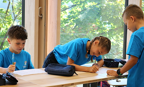 Ein Mädchen steht an einem Schultisch und schreibt auf ein Blatt Papier. Vor ihr steht ein Junge, neben ihr sitzt ein Junge.