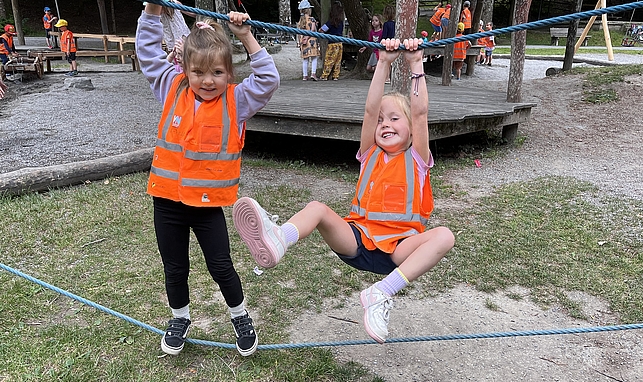 Zwei Mädchen spielen an einer Klettermöglichkeit. Ein Mädchen steht auf einem Seil und hält sich an einem Seil über ihrem Kopf fest, das zweite Mädchen hält sich am oberen Seil fest und baumelt mit den Füßen in der Luft. 