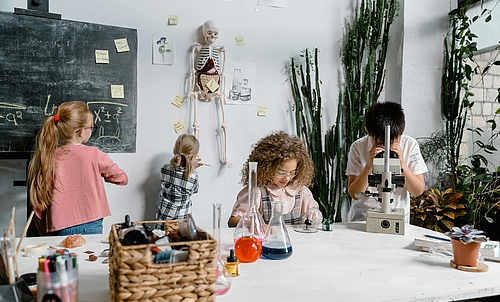 Ein Junge schaut durch ein Mikroskop, ein Mädchen experimentiert mit einer Pipette, ein Mädchen schreibt auf eine Tafel und ein Mädchen begutachtet ein Skelett.