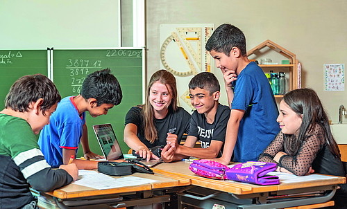 Drei Kinder und eine Lehrerin sitzen an Tischen, zwei Kinder stehen. Die Lehrerin klickt auf einem Laptop und zeigt den Kindern etwas.