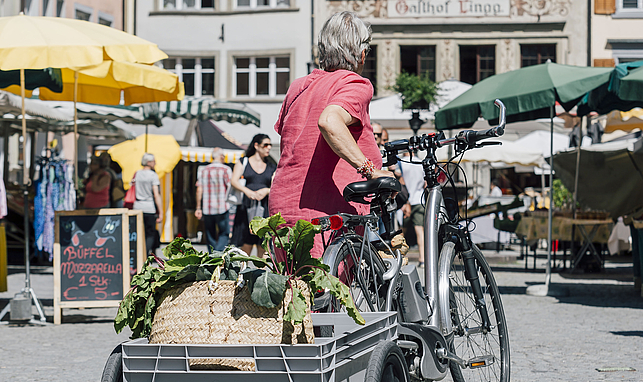 Eine ältere Dame schiebt ein Elektrofahrrad mit kleinem Anhänger über den Marktplatz in Feldkirch. Im Hintergrund sind Marktstände. 