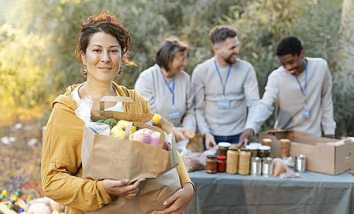 Eine Dame hält eine Tüte mit Lebensmittel in den Händen. Hinter ihr stehen drei Personen an einem Tisch, auf dem noch mehr Lebensmittel stehen. 